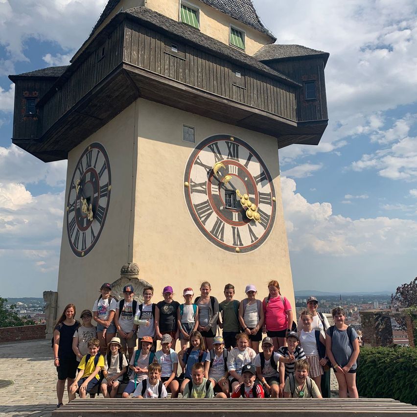 Eine Gruppe von Kindern und Erwachsenen posiert für ein Foto vor einem Uhrturm mit einem großen Zifferblatt an einem sonnigen Tag.