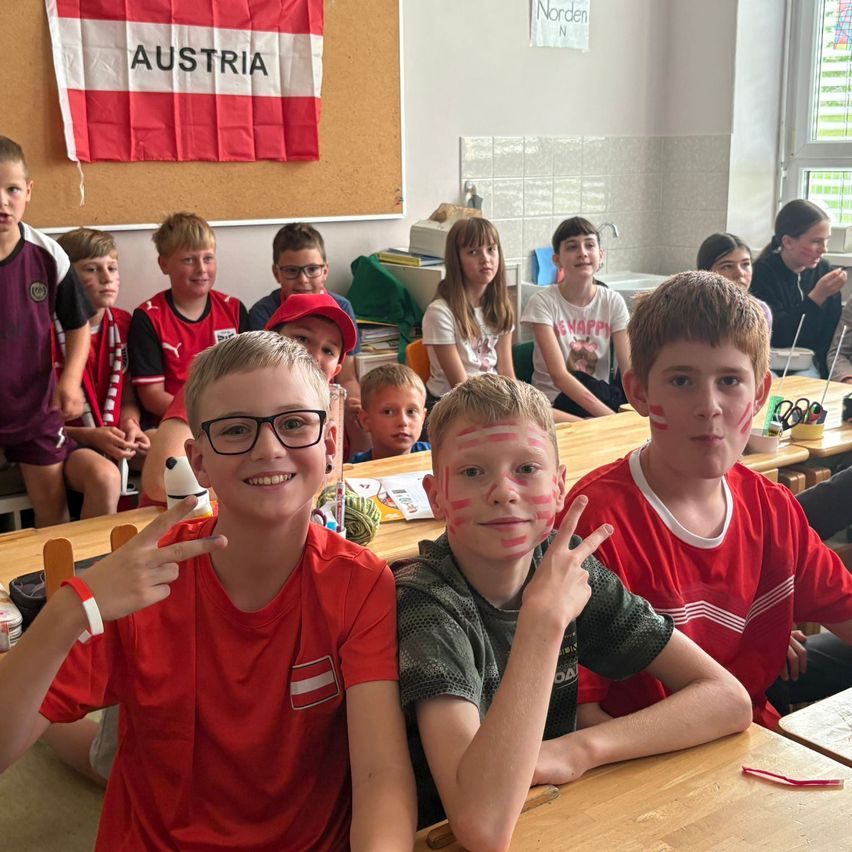 Eine Gruppe von Kindern mit roten Trikots, auf denen die Flagge von Österreich gemalt ist, sitzen an Tischen in einem Klassenzimmer. Die Flagge von Österreich hängt an der Wand hinter ihnen.