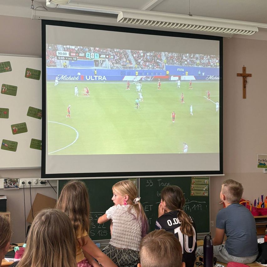 In einem Klassenzimmer sitzen Kinder vor einer Projektionsleinwand, die ein Fußballspiel zeigt. Das Spiel steht 2 zu 1. Ein Kreuz schmückt die Wand.