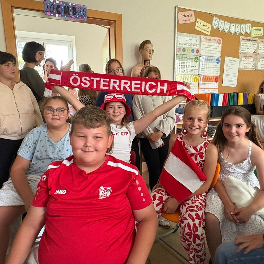 Eine Gruppe von Kindern mit roten und weißen Schals hält ein Banner mit der Aufschrift Osterreich hoch, sie scheinen sich in einem Klassenzimmer zu befinden.