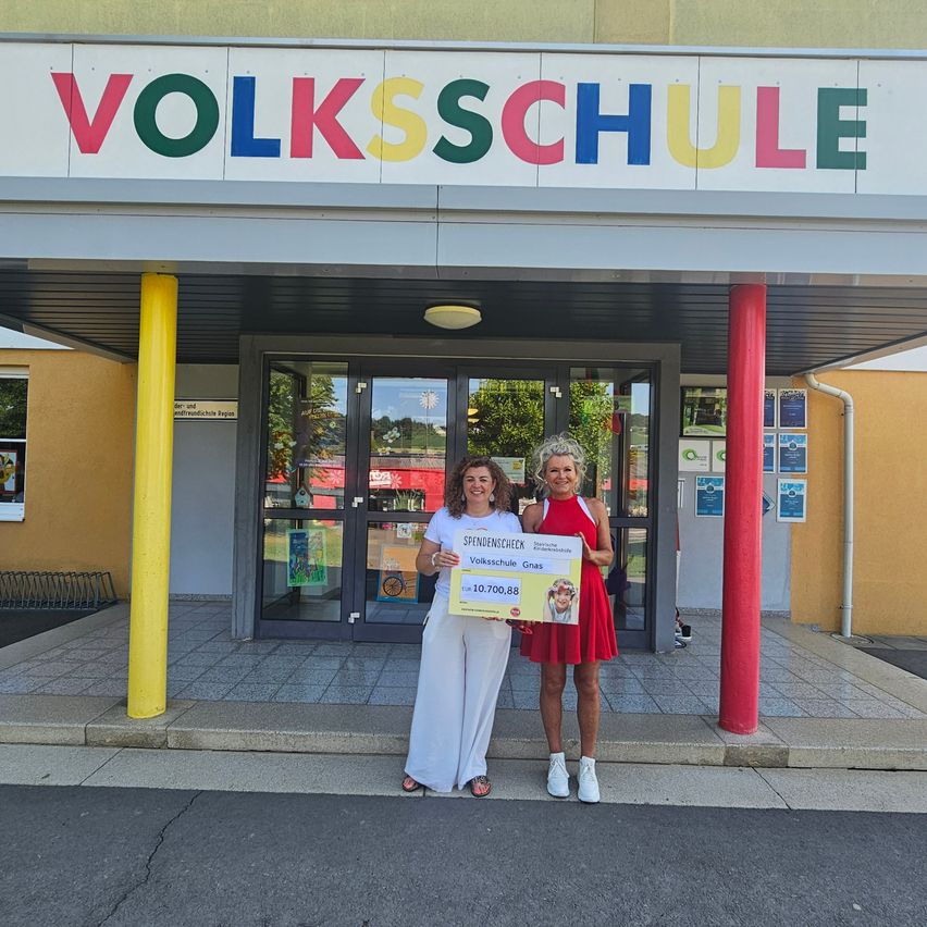 Zwei Frauen stehen vor einem Gebäude namens Volksschule und halten einen Scheck über 10.700,88 Euro. Die Frau links trägt ein weißes Outfit, die andere ein rotes Kleid.