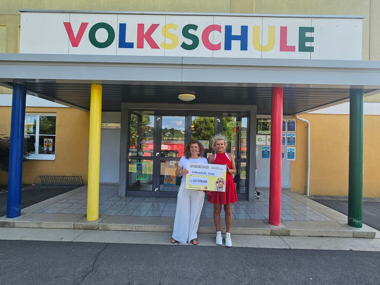Zwei Frauen stehen vor einem Gebäude namens Volksschule und halten einen Scheck über 10.700,88 Euro. Die Frau links trägt ein weißes Outfit, die andere ein rotes Kleid.