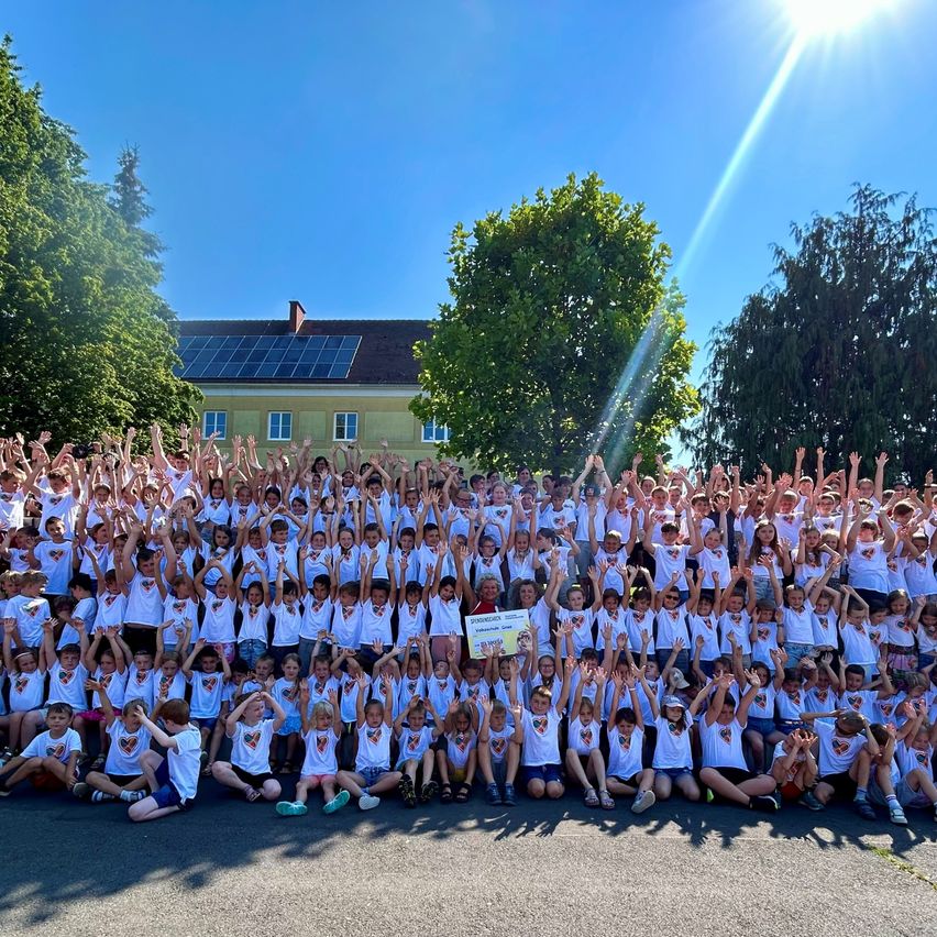 Eine große Gruppe von Kindern in identischen weißen T-Shirts posiert für ein Foto mit erhobenen Händen vor einem Gebäude mit Solarmodulen auf dem Dach.