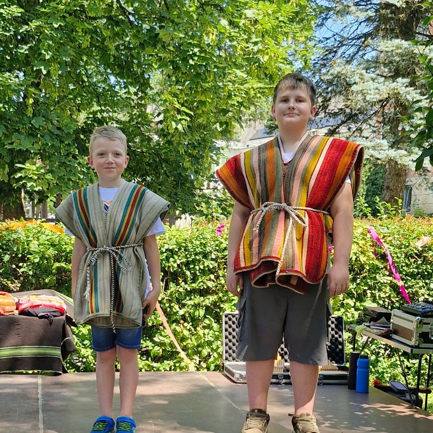 Zwei Jungen stehen auf einer Bühne und tragen bunte, selbstgemachte Westen. Sie lächeln, und hinter ihnen sind grüne Pflanzen und Gegenstände.