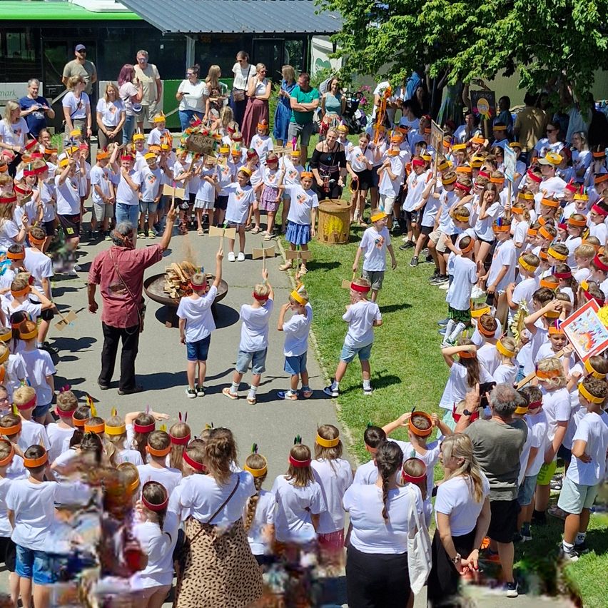 Eine Gruppe von Kindern in weißen Shirts und orangefarbenen Stirnbändern steht im Kreis. Ein Mann steht vorne und hält einen Trommel. Zuschauer beobachten von den Seiten. Das Gebiet ist von Bäumen und Grünflächen umgeben.