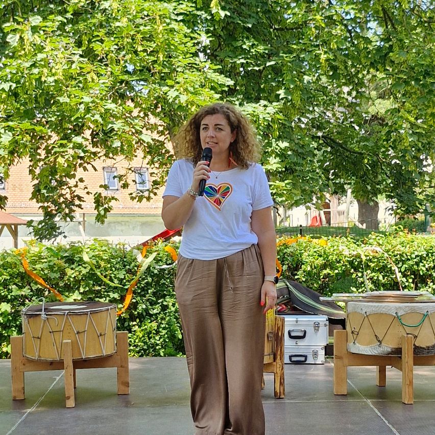 Eine Frau steht auf der Bühne und hält ein Mikrofon in der Hand. Sie trägt ein weißes T-Shirt mit einem farbenfrohen Herzdesign. Um sie herum stehen zwei Trommeln auf hölzernen Stühlen und eine Kulisse aus Bäumen und Grün.