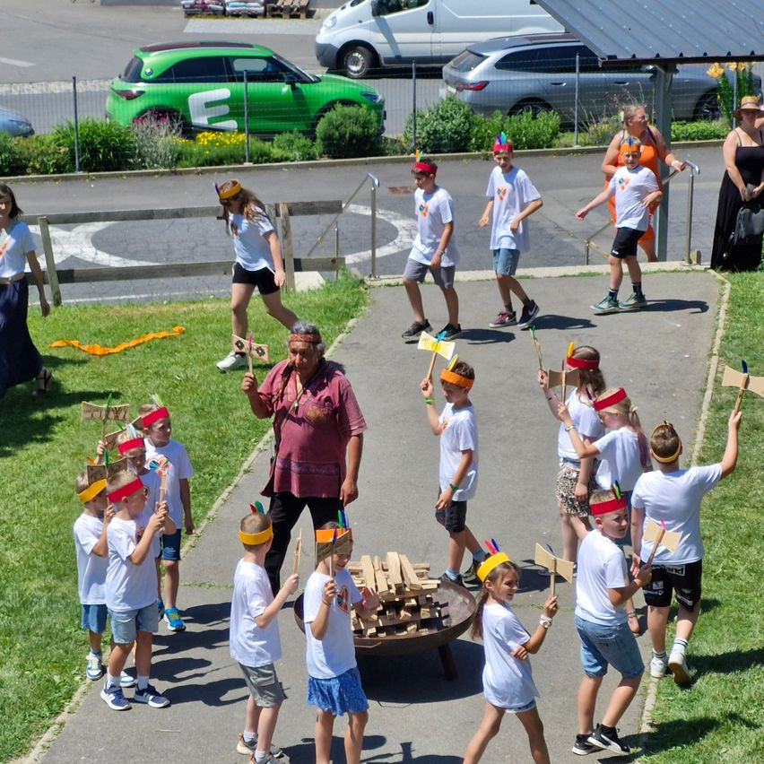 Eine Gruppe von Kindern mit Federkopfbändern, die in einer Reihe gehen, Stöcke halten und einem Mann vor ihnen. Autos im Hintergrund geparkt.