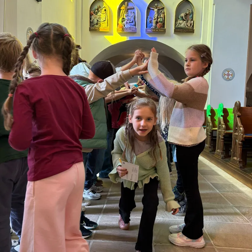 Eine Gruppe von Kindern in einer Kirche, eine hält ein Papier, eine andere hält die Hände einer Mädchen in einem gestreiften Pullover.