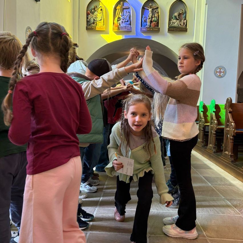 Eine Gruppe von Kindern in einer Kirche, eine hält ein Papier, eine andere hält die Hände einer Mädchen in einem gestreiften Pullover.