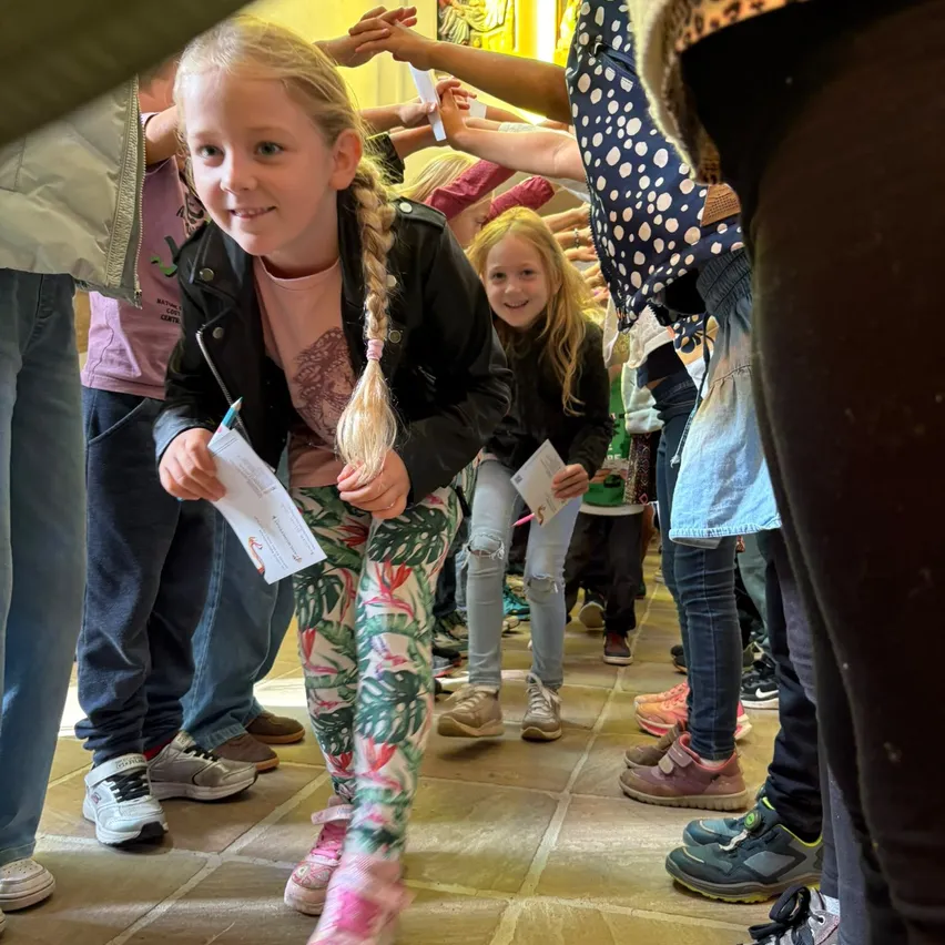 Ein junges Mädchen mit geflochtenen Haaren steht in einer Reihe, hält ein Stück Papier in der Hand und lächelt. Sie ist von anderen Kindern und Erwachsenen umgeben, die alle Freizeitkleidung tragen.