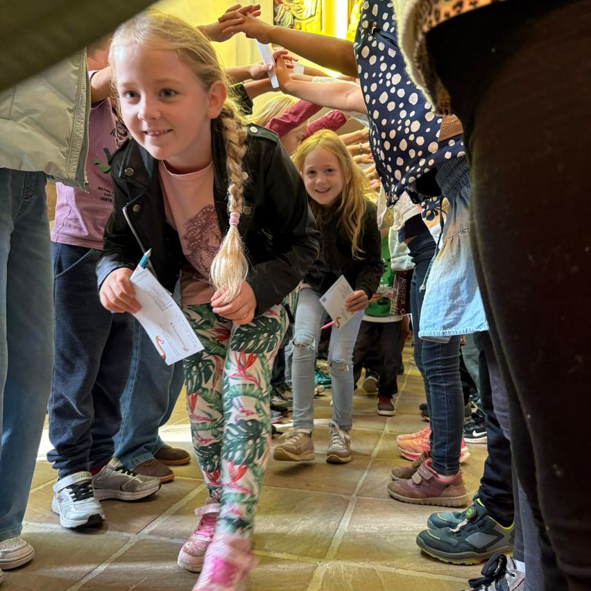 Ein junges Mädchen mit geflochtenen Haaren steht in einer Reihe, hält ein Stück Papier in der Hand und lächelt. Sie ist von anderen Kindern und Erwachsenen umgeben, die alle Freizeitkleidung tragen.