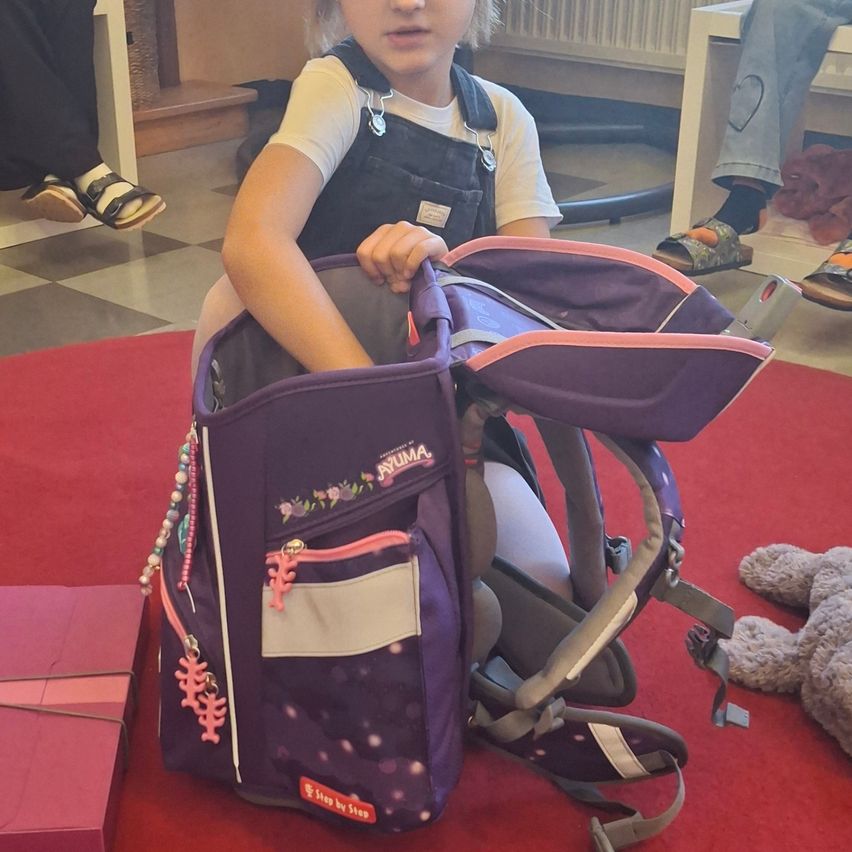 Ein kleines Mädchen sitzt auf einem roten Teppich und hält einen lila Rucksack mit rosa Akzenten. Sie trägt ein weißes Hemd und eine schwarze Schürze. Ein rosa Kasten und ein Plüschtier sind in der Nähe.