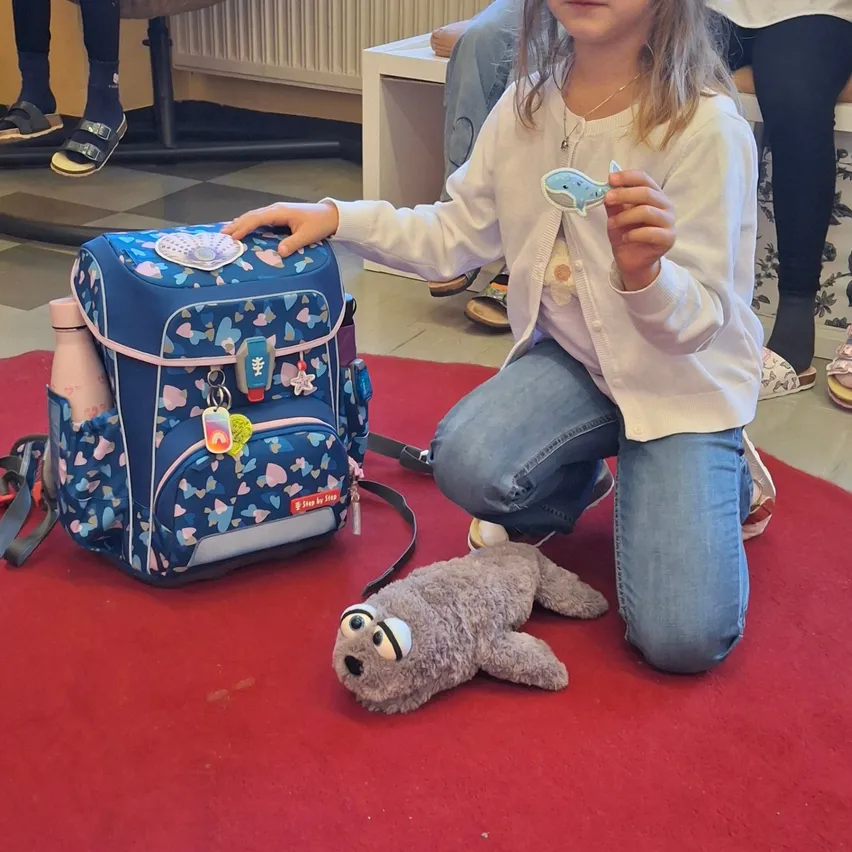 Ein kleines Mädchen kniet auf einem roten Teppich und hält einen blauen Rucksack und einen Wal-Schlüsselanhänger. Sie trägt eine weiße Strickjacke und Jeans.