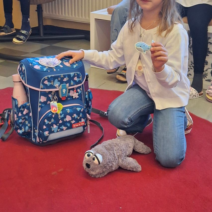 Ein kleines Mädchen kniet auf einem roten Teppich und hält einen blauen Rucksack und einen Wal-Schlüsselanhänger. Sie trägt eine weiße Strickjacke und Jeans.