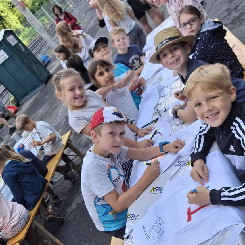 Eine Gruppe von Kindern sitzt an einem Tisch und zeichnet auf weiße Papiere. Einige tragen Hüte und Brillen. Hinter ihnen sind andere Kinder und Erwachsene, möglicherweise Lehrer. Links ist eine grüne mobile Toilette zu sehen.