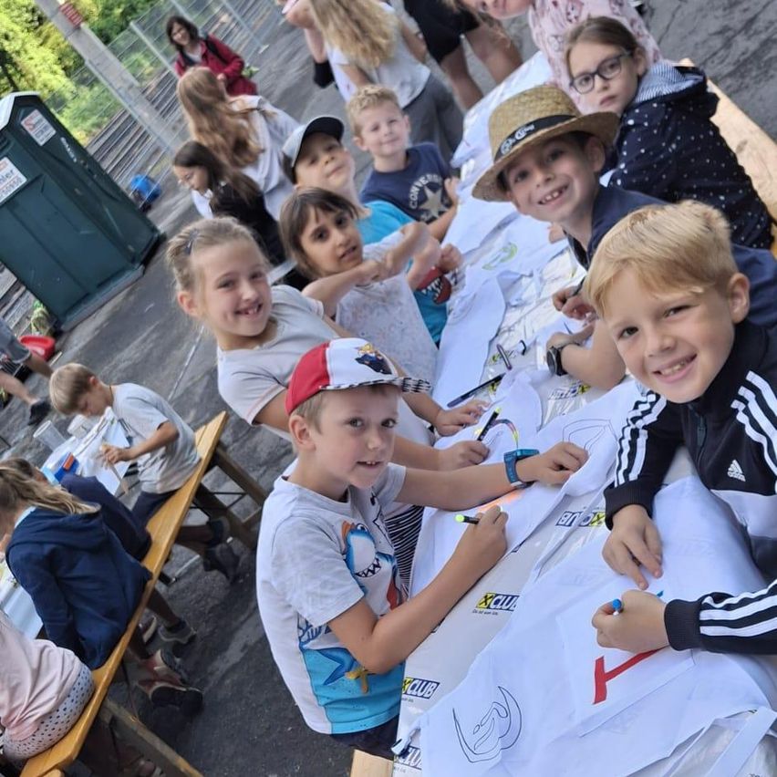 Eine Gruppe von Kindern sitzt an einem Tisch und zeichnet auf weiße Papiere. Einige tragen Hüte und Brillen. Hinter ihnen sind andere Kinder und Erwachsene, möglicherweise Lehrer. Links ist eine grüne mobile Toilette zu sehen.