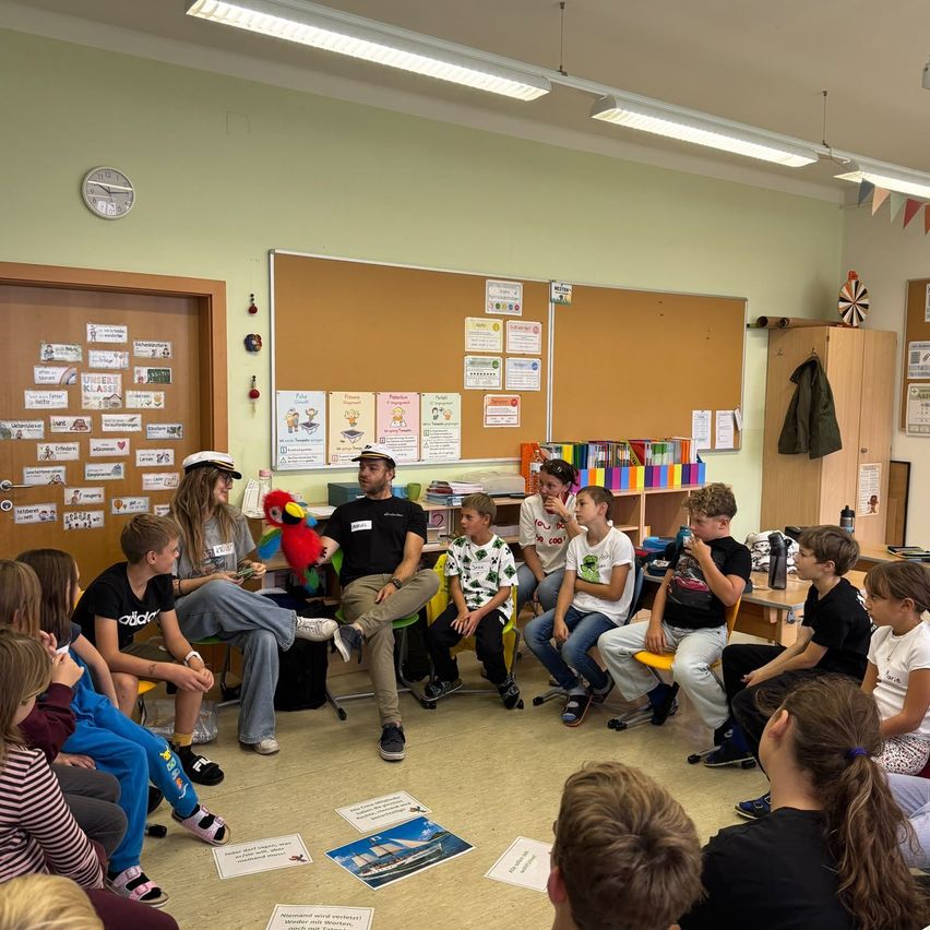 Eine Gruppe von Kindern und zwei Erwachsenen sitzen im Kreis in einem Klassenzimmer. Ein Erwachsener hält einen Papagei und die Kinder hören aufmerksam zu. Papiere sind auf dem Boden verstreut.