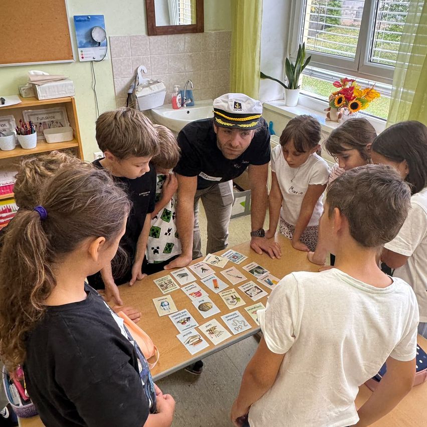 Eine Gruppe von Kindern und einem Erwachsenen versammelt sich um einen Tisch mit Karten in einem Klassenzimmer. Der Erwachsene trägt einen Hut und erklärt den Kindern etwas.