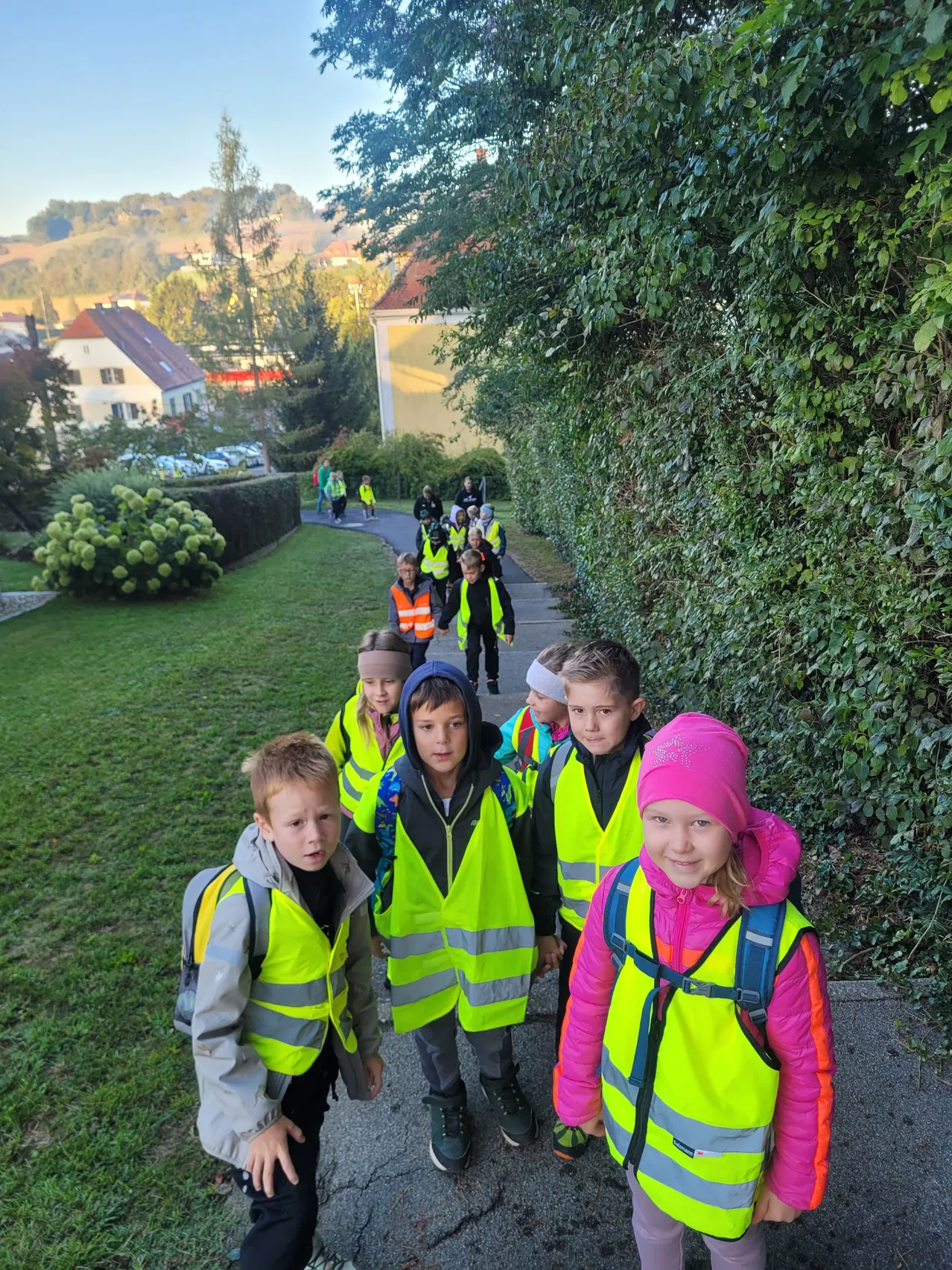Eine Gruppe von Kindern in neongelben Westen geht auf einem Bürgersteig mit einer Hecke an der Seite.