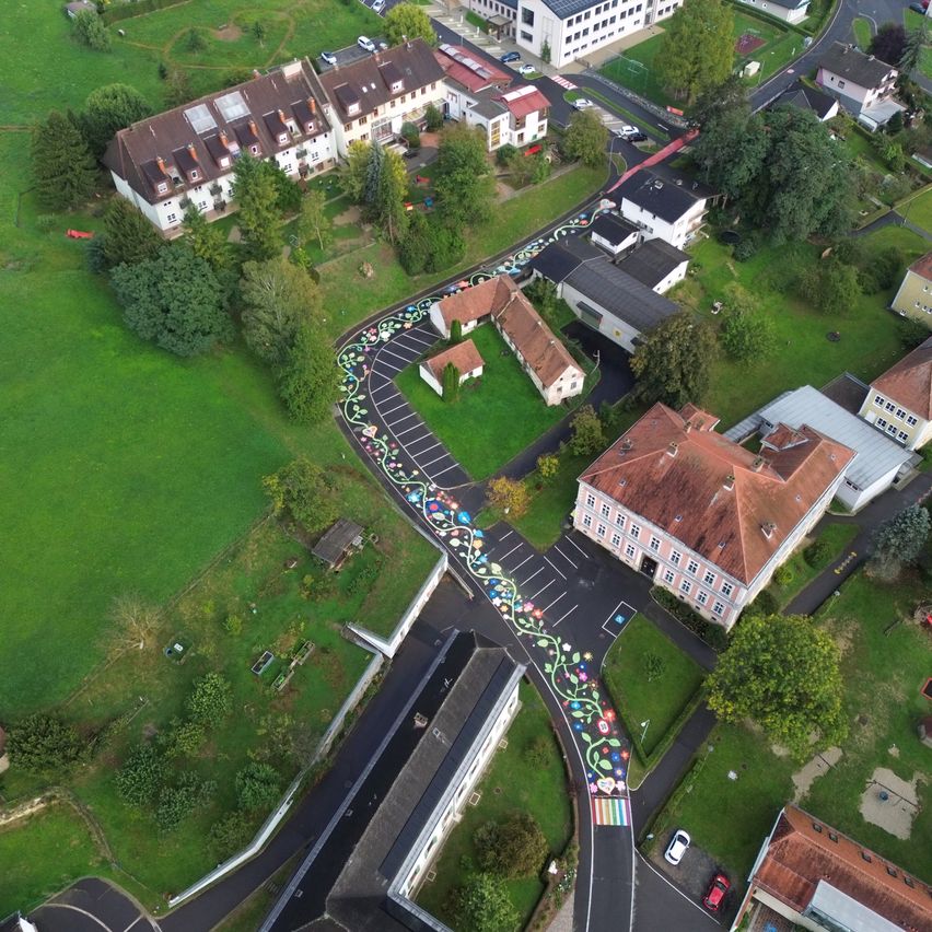 Ein Luftbild eines Wohngebiets mit einem farbenfrohen Straßendesign. Die Straße ist mit Blumen verziert und hat ein Regenbogenmuster.