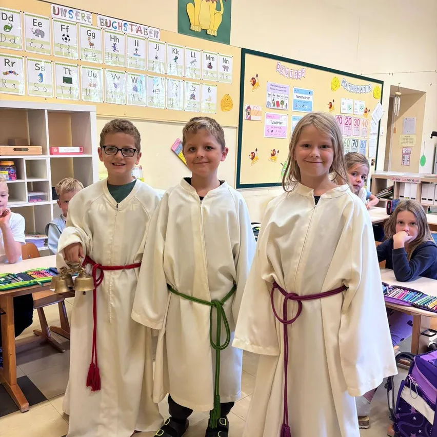 Drei Kinder in weißen Roben mit roten und grünen Schärpen stehen in einem Klassenzimmer.