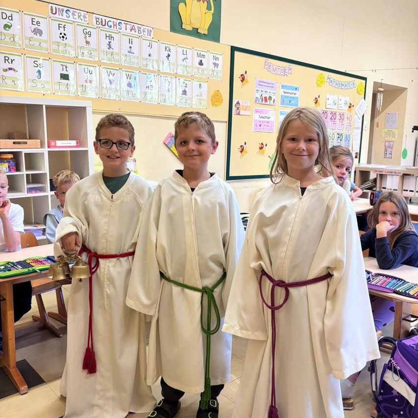 Drei Kinder in weißen Roben mit roten und grünen Schärpen stehen in einem Klassenzimmer.
