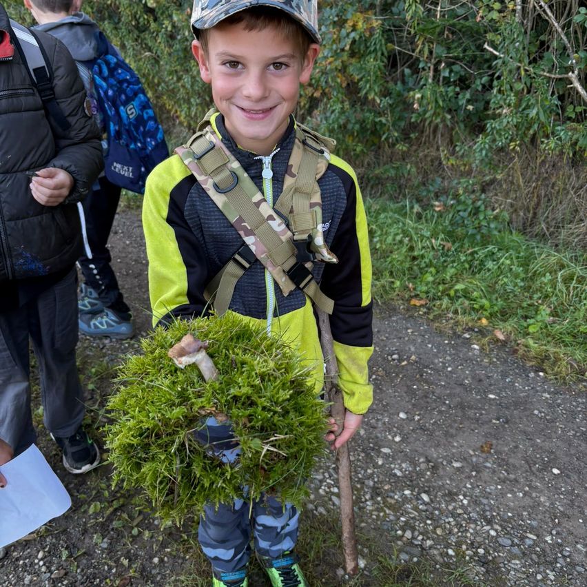 Ein junger Junge steht auf einem Kiesweg, lächelt und hält einen Stock mit einem Haufen Moos und einem Pilz daran.