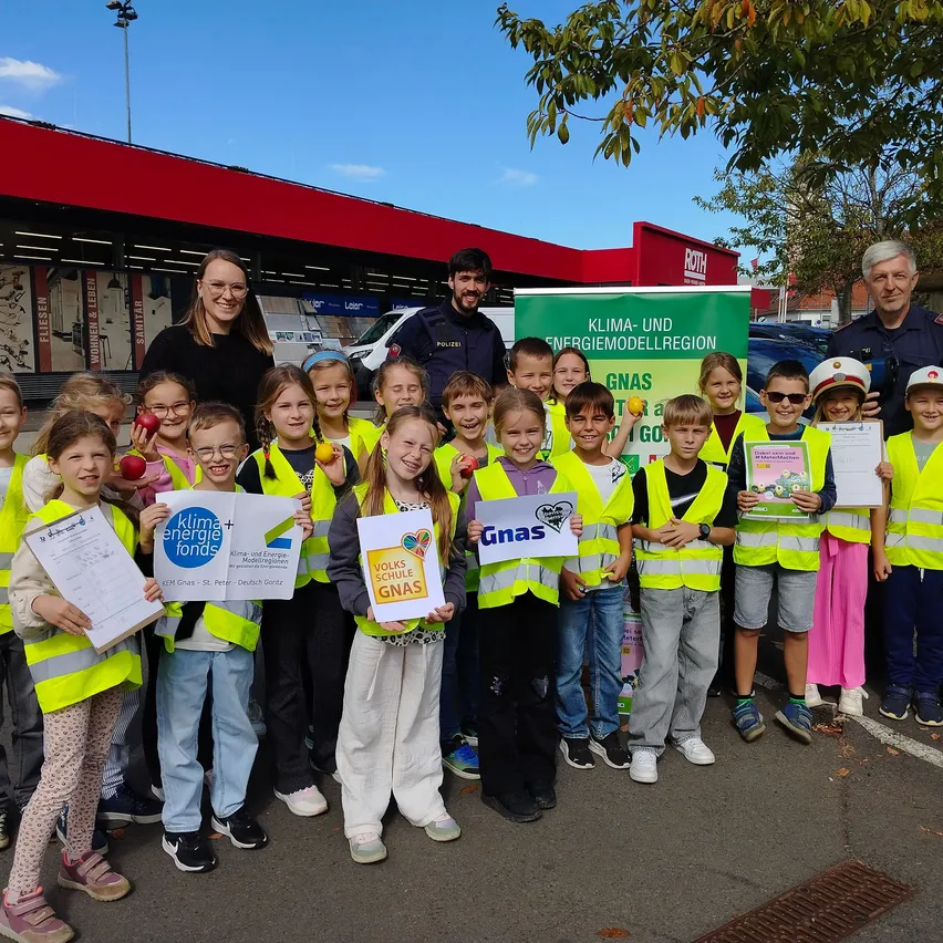 Eine Gruppe von Kindern und Erwachsenen steht vor einem Gebäude mit einem grünen Banner, das Klima- und Energiemodellregion GNAS liest. Die Kinder tragen gelbe Westen und halten Urkunden.