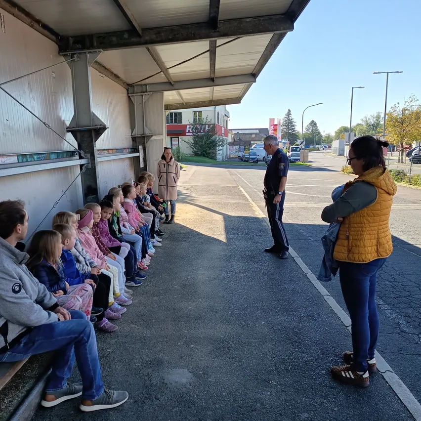 Eine Gruppe von Kindern und Erwachsenen sitzt auf einer Bank unter einem Schutzdach, ein Polizist und eine Frau stehen in der Nähe.