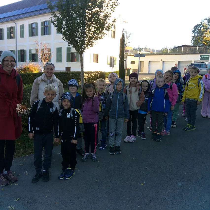 Eine Gruppe von Kindern und zwei Erwachsenen stehen vor einem Gebäude. Die Kinder tragen Jacken und Turnschuhe.