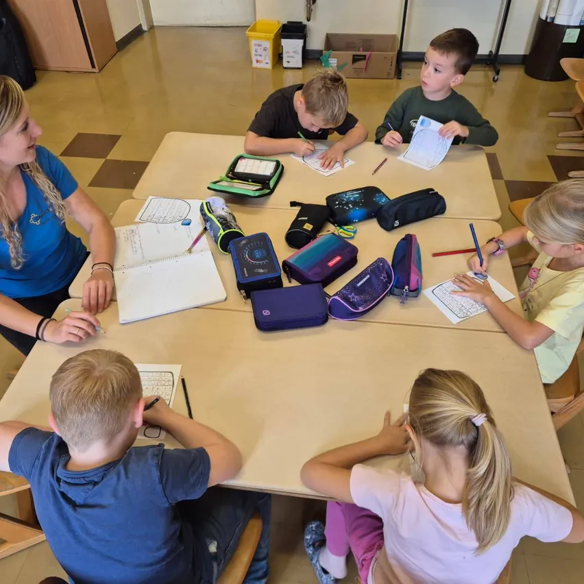 Eine Gruppe von Kindern sitzt in einem Klassenzimmer um einen Tisch herum und schreibt mit einer Lehrerin.