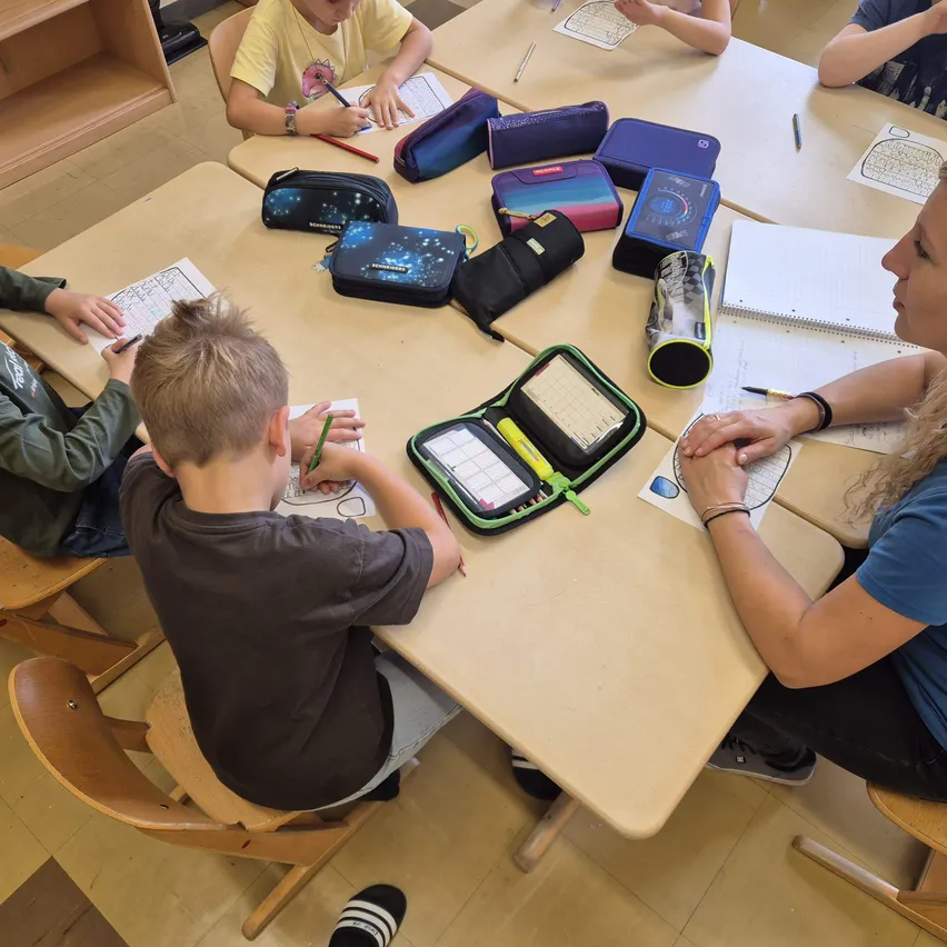 Eine Klassenzimmerszene mit einer Lehrerin und vier jungen Jungen, die an einem Tisch sitzen und Schreibaktivitäten durchführen. Die Lehrerin schaut den Jungen auf ihrer rechten Seite an, während die anderen Jungen auf ihre eigenen Aufgaben konzentriert sind.