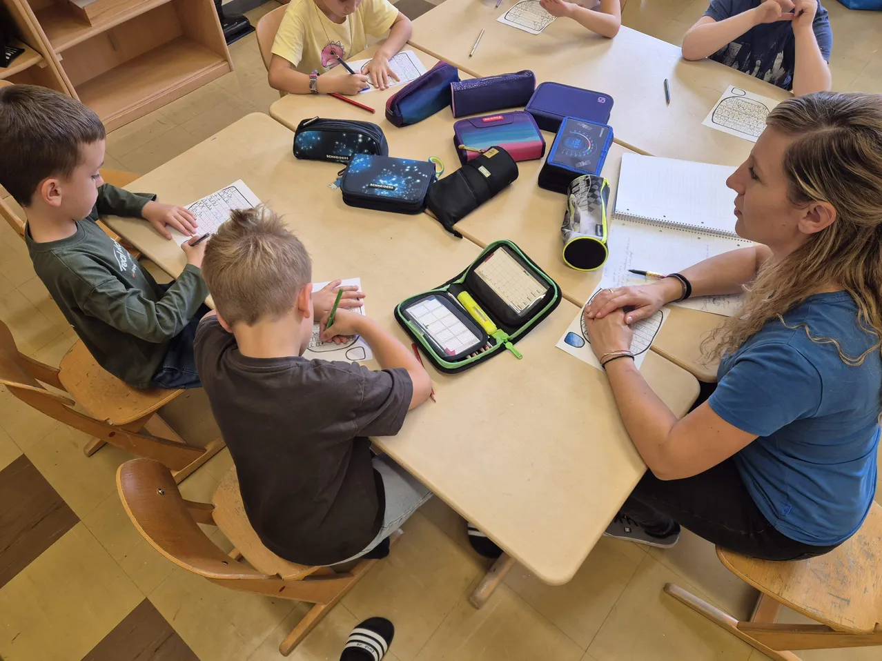 Eine Klassenzimmerszene mit einer Lehrerin und vier jungen Jungen, die an einem Tisch sitzen und Schreibaktivitäten durchführen. Die Lehrerin schaut den Jungen auf ihrer rechten Seite an, während die anderen Jungen auf ihre eigenen Aufgaben konzentriert sind.