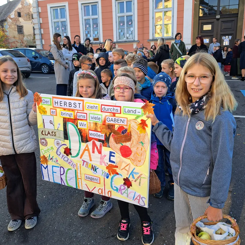Drei junge Mädchen halten ein farbenfrohes Schild mit Wörtern in mehreren Sprachen, vor einer Menschenmenge aus Kindern und Erwachsenen. Das Schild hat Zeichnungen von Obst und Gemüse. Die Kinder tragen Winterkleidung und halten Körbe.
