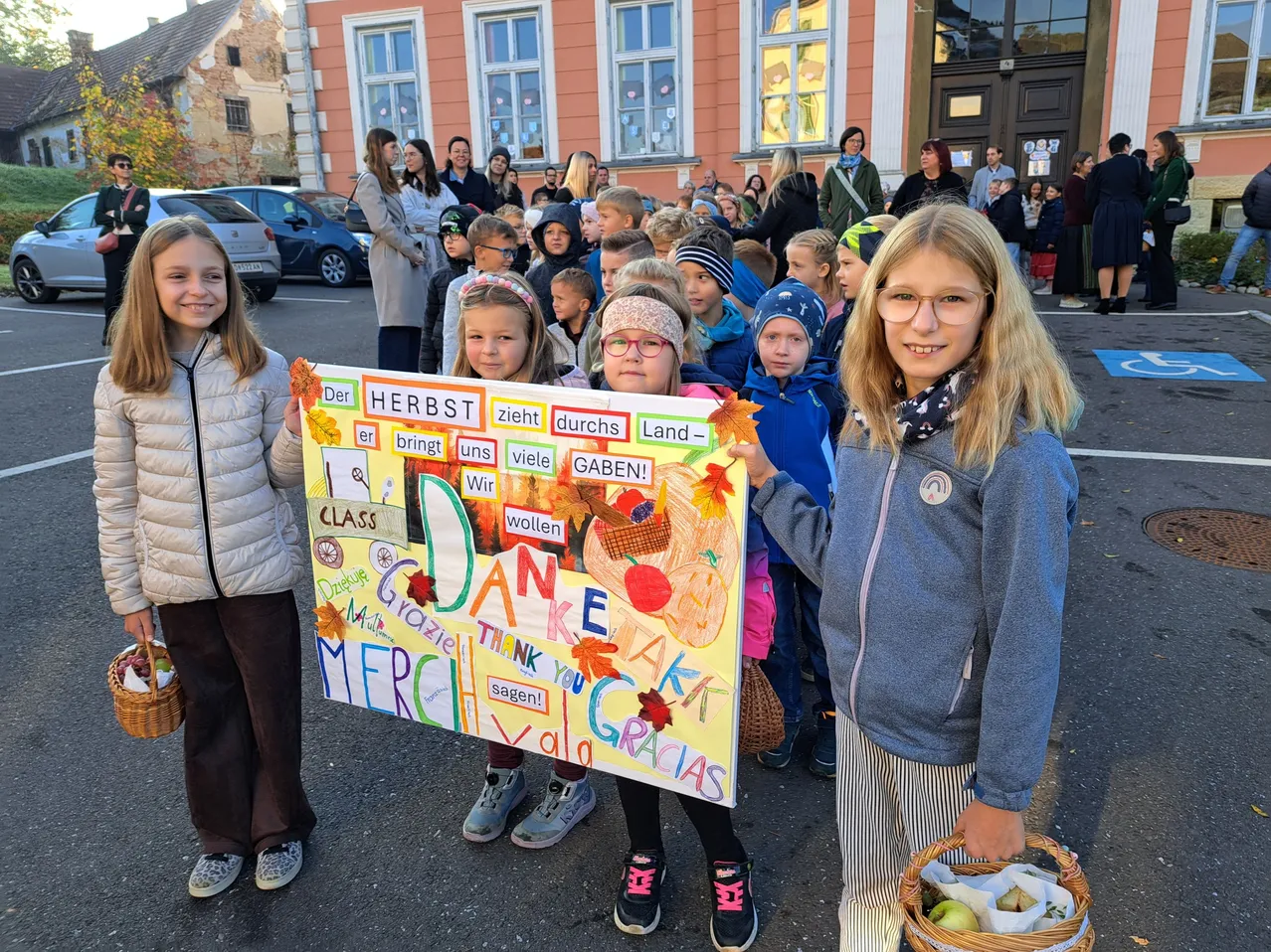 Drei junge Mädchen halten ein farbenfrohes Schild mit Wörtern in mehreren Sprachen, vor einer Menschenmenge aus Kindern und Erwachsenen. Das Schild hat Zeichnungen von Obst und Gemüse. Die Kinder tragen Winterkleidung und halten Körbe.
