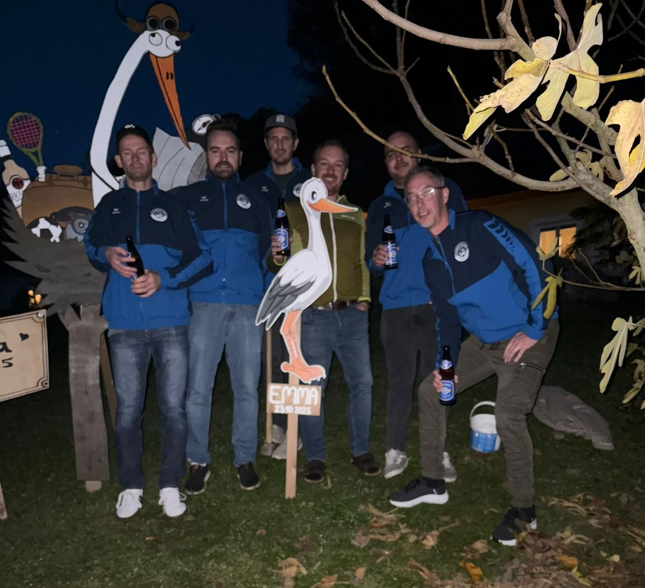 Sechs Männer posieren nachts für ein Foto, alle in blauen Jacken, vor einem Schild mit der Aufschrift Emma. Sie halten jeweils eine Flasche Bier in der Hand. Dahinter steht ein großer Storch und ein Baum mit gelben Blättern.