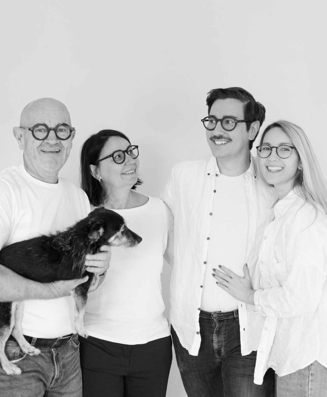 Ein monochromes Foto zeigt eine Familie aus vier Personen, bestehend aus zwei Männern und zwei Frauen, die eng beieinanderstehen. Der Mann auf der linken Seite hält einen nassen Hund, während die anderen drei lächeln. Sie tragen alle weiße Hemden, und der Mann auf der rechten Seite trägt eine Jeans.