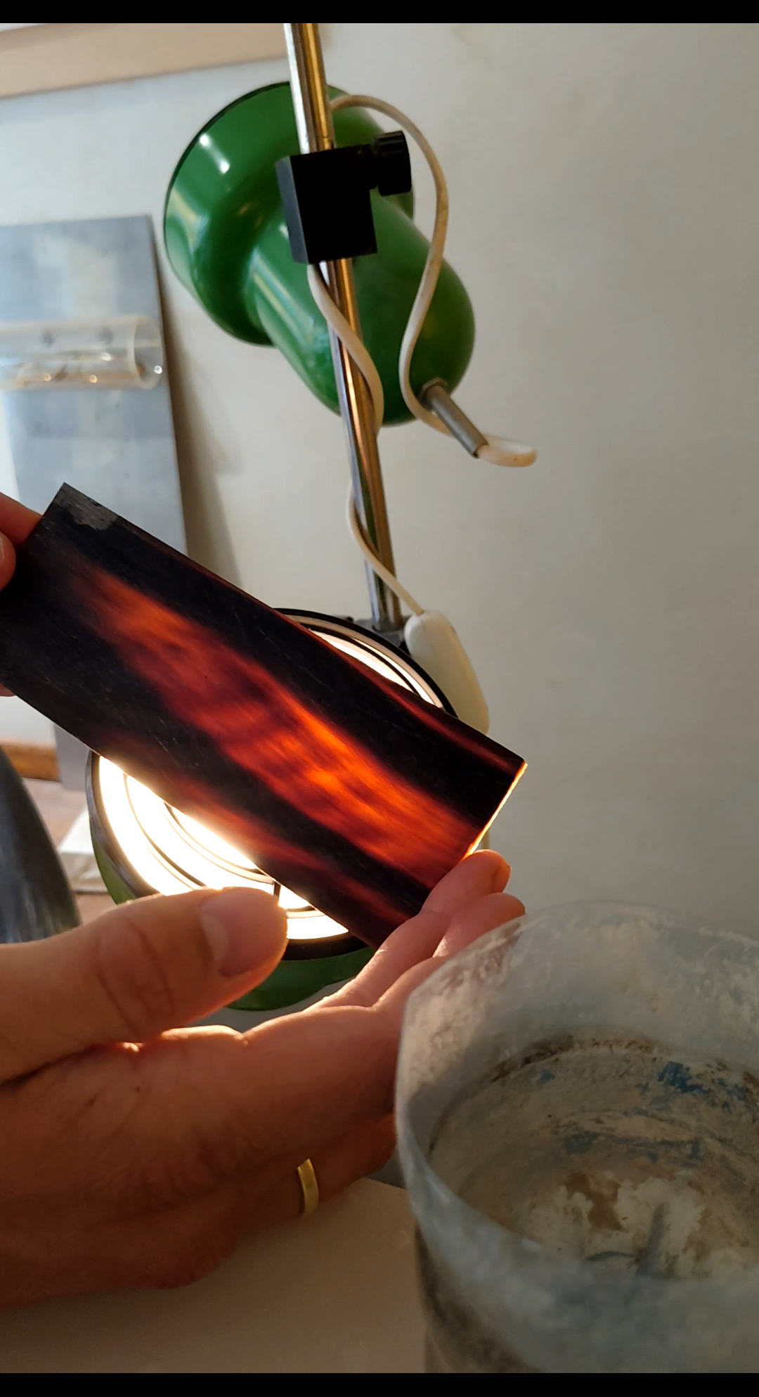 A person holds a piece of wood with a flame-like pattern under a green desk lamp.