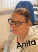 Eine Frau mit Brille sitzt vor einer weißen Wand und trägt ein weißes Hemd und eine beige Jacke. Der Name 'Anita' steht über ihrem Kopf an der Wand. Hinter ihr befindet sich ein blauer Stuhl und ein weißer Heizkörper.