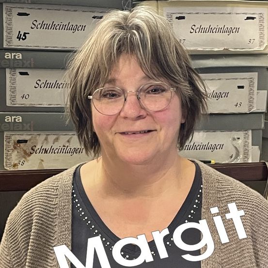 Eine Frau mit kurzen Haaren und Brille steht vor einer Wand mit verschiedenen Lagerboxen. Die Frau lächelt und schaut direkt in die Kamera. Die Boxen haben Etiketten mit den Aufschriften 'Schuheinlagen' und Zahlen wie 45, 37, 40, 43 und 38.