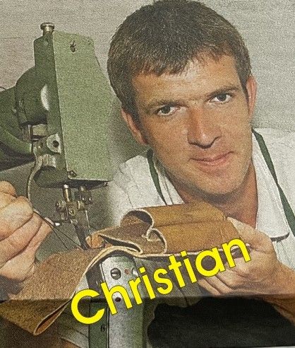 Ein Mann in einem weißen Hemd und grüner Schürze hält ein Stück braunes Leder in der Nähe einer Maschine mit dem Wort Christian darauf.