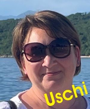 Eine Frau mit kurzen Haaren und Sonnenbrille lächelt in die Kamera. Der Name Uschi ist in gelbem Text zu sehen. Im Hintergrund befindet sich eine ruhige Wasserfläche mit Bergen in der Ferne.
