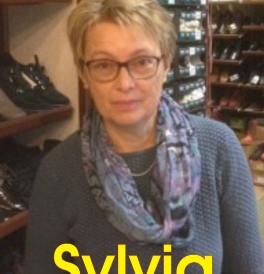 Eine Frau mit kurzen blonden Haaren, die eine Brille und einen Schal trägt, steht vor einem Schuhgeschäft mit Regalen voller Schuhe.