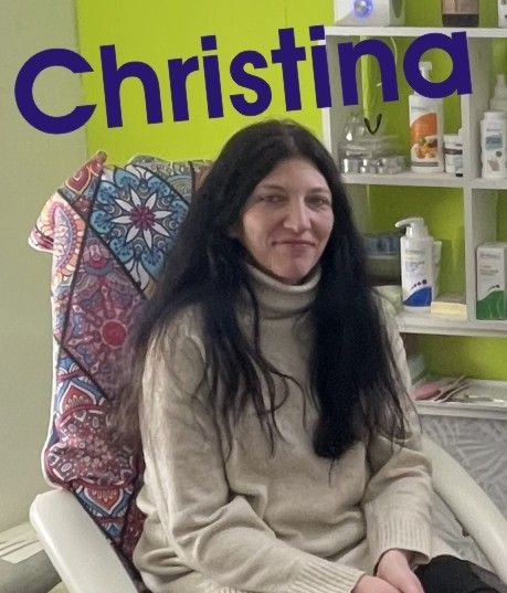 Eine Frau mit langen dunklen Haaren sitzt in einem Stuhl mit einem bunten Kissen hinter ihr. Sie trägt ein beigefarbenes Rollkragenshirt und steht vor einer grünen Wand mit Regalen voller Produkte.