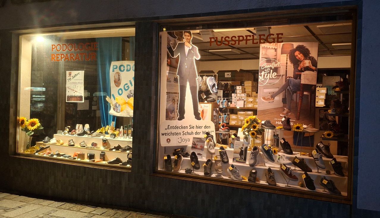Ladenfenster mit einer Ausstellung von Joya-Schuhen. Eine Ausschnitt eines Mannes in einem Anzug ist auf dem Fenster. Das Fenster zeigt verschiedene Schuhe und Sonnenblumen.