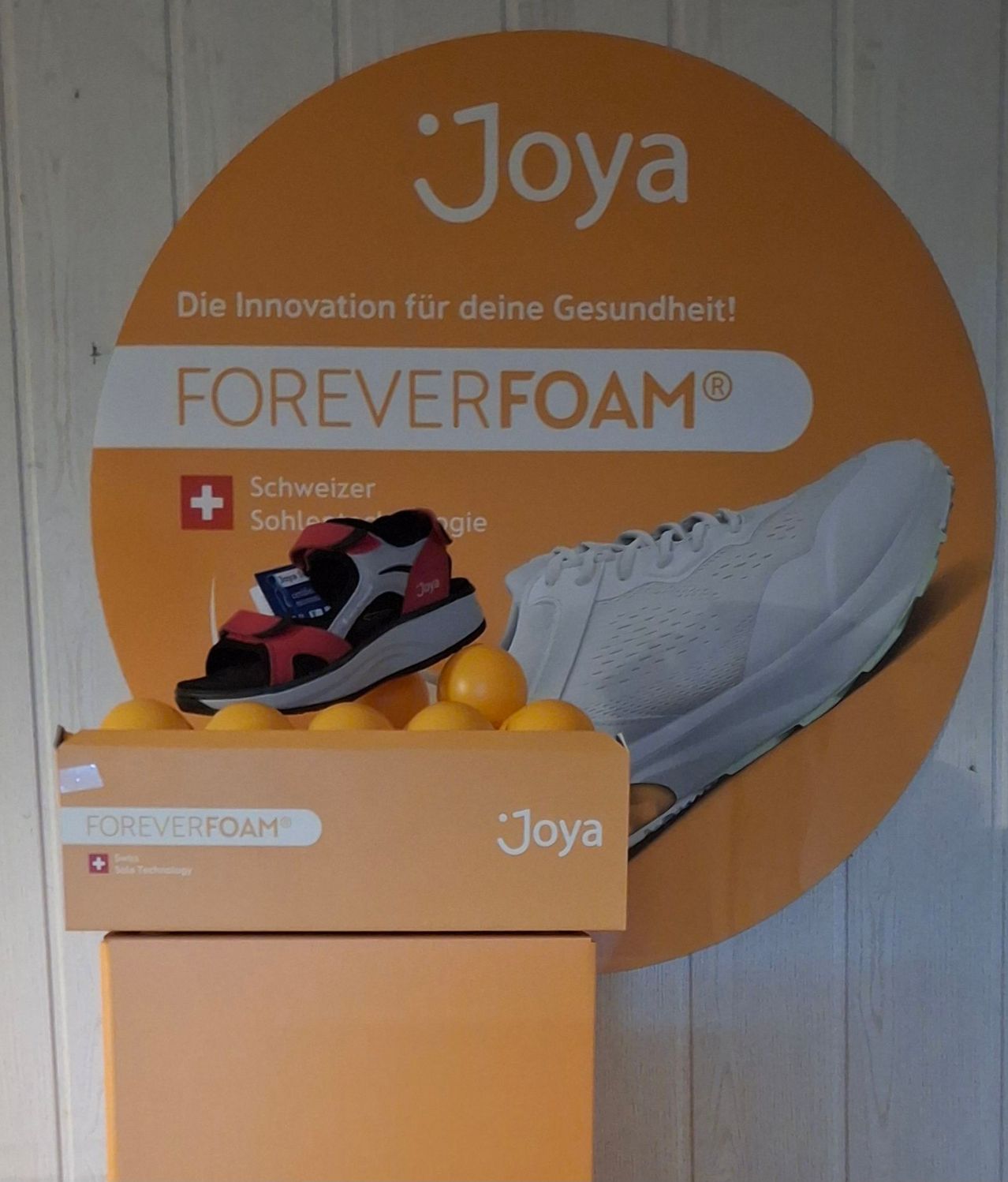 Ein orangefarbenes Plakat mit dem Joya-Logo und dem Slogan 'ForeverFoam'. Unter dem Logo steht eine Schachtel Joya-Schuhe auf einem Display, begleitet von einem Stapel Orangen.