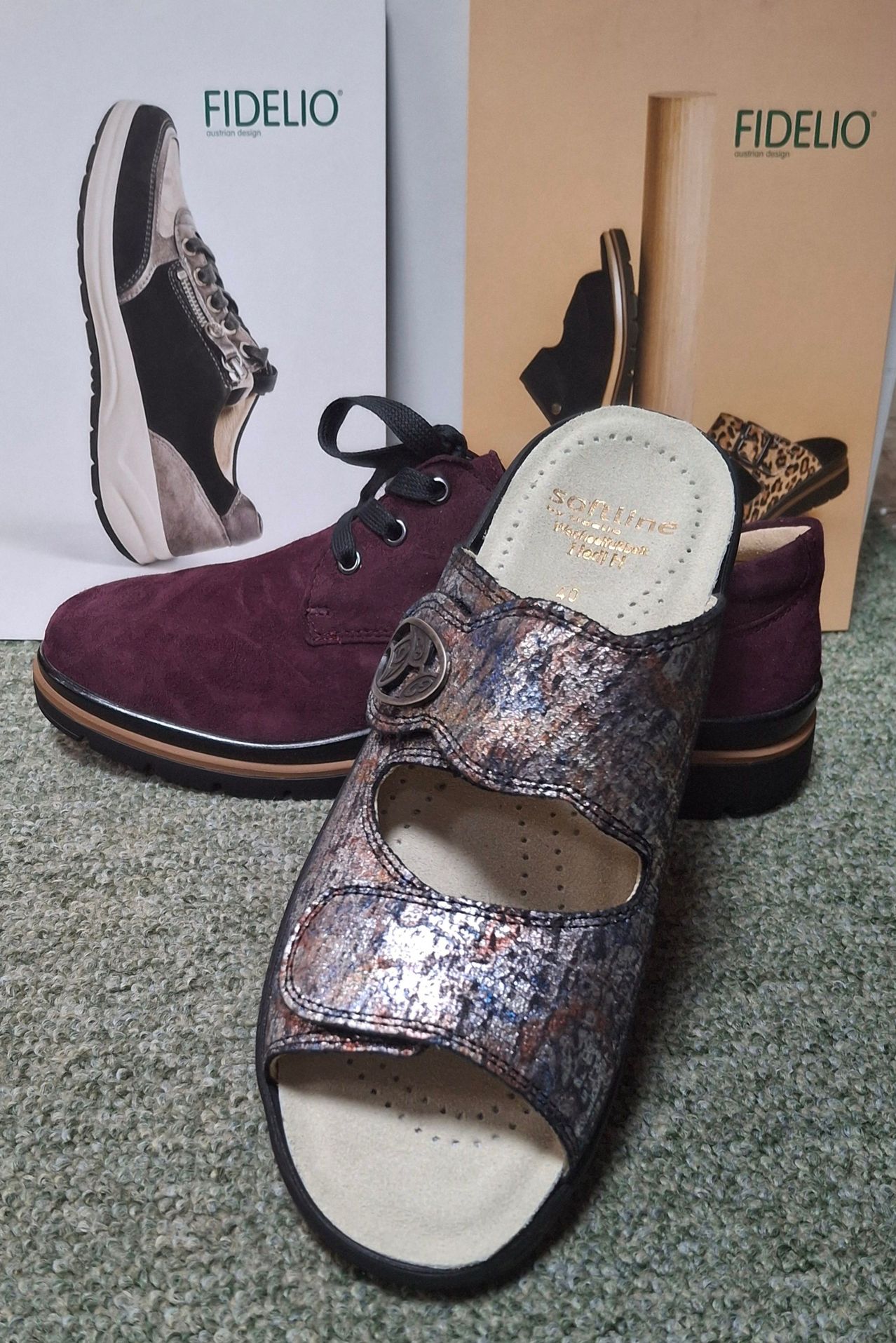 Zwei Paar Schuhe liegen auf einem Teppichboden. Ein Paar sind bordeauxrote Wildlederstiefel mit Schnürsenkeln, und das andere sind silberne Metallic-Sandalen mit einer Schnalle. Dahinter befindet sich eine Schuhschachtel mit Leopardenmuster-Schlappen.