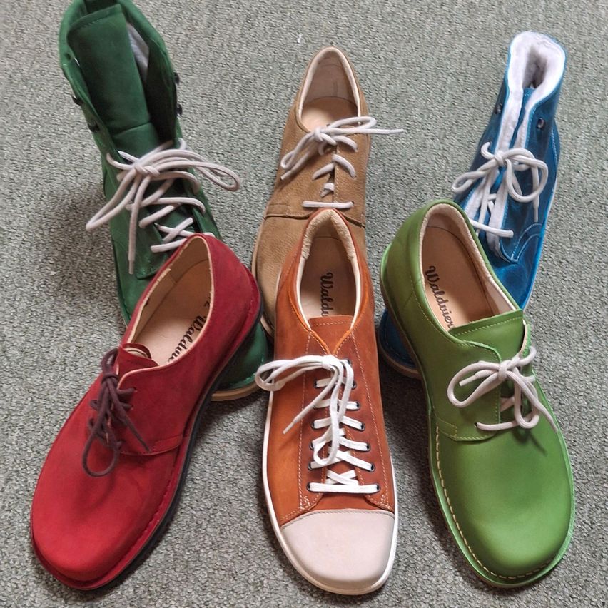 Fünf Paar Schuhe in verschiedenen Farben und Stilen sind auf einem grauen Teppich ausgestellt. Ein roter Schuh, ein brauner Schuh, ein blauer Schuh, ein grüner Schuh und ein grüner Stiefel sind aufgereiht.