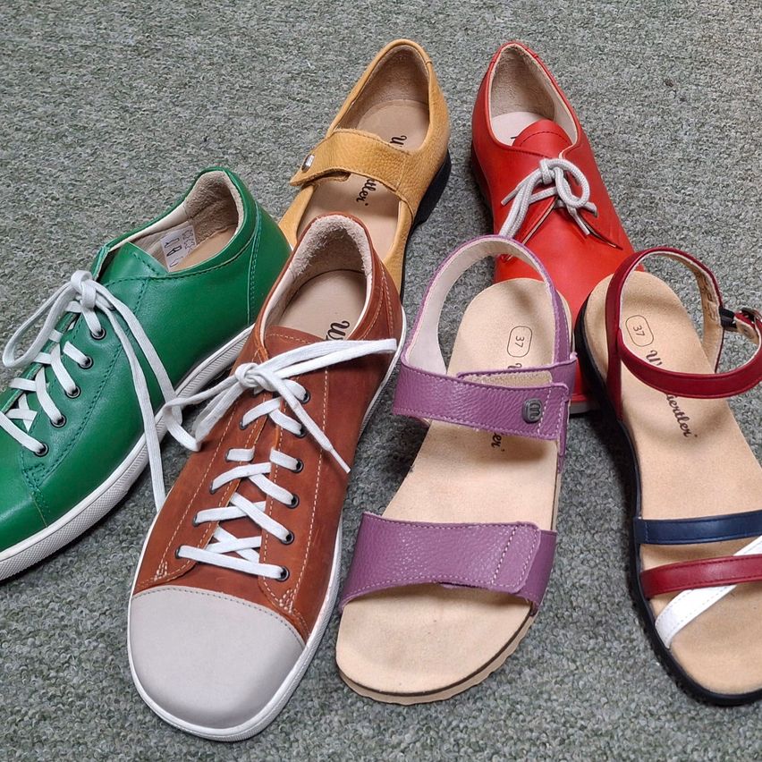 Fünf Paar Schuhe in verschiedenen Farben liegen auf einem Teppich. Sie umfassen grüne, braune, gelbe, rote und lila Schuhe.