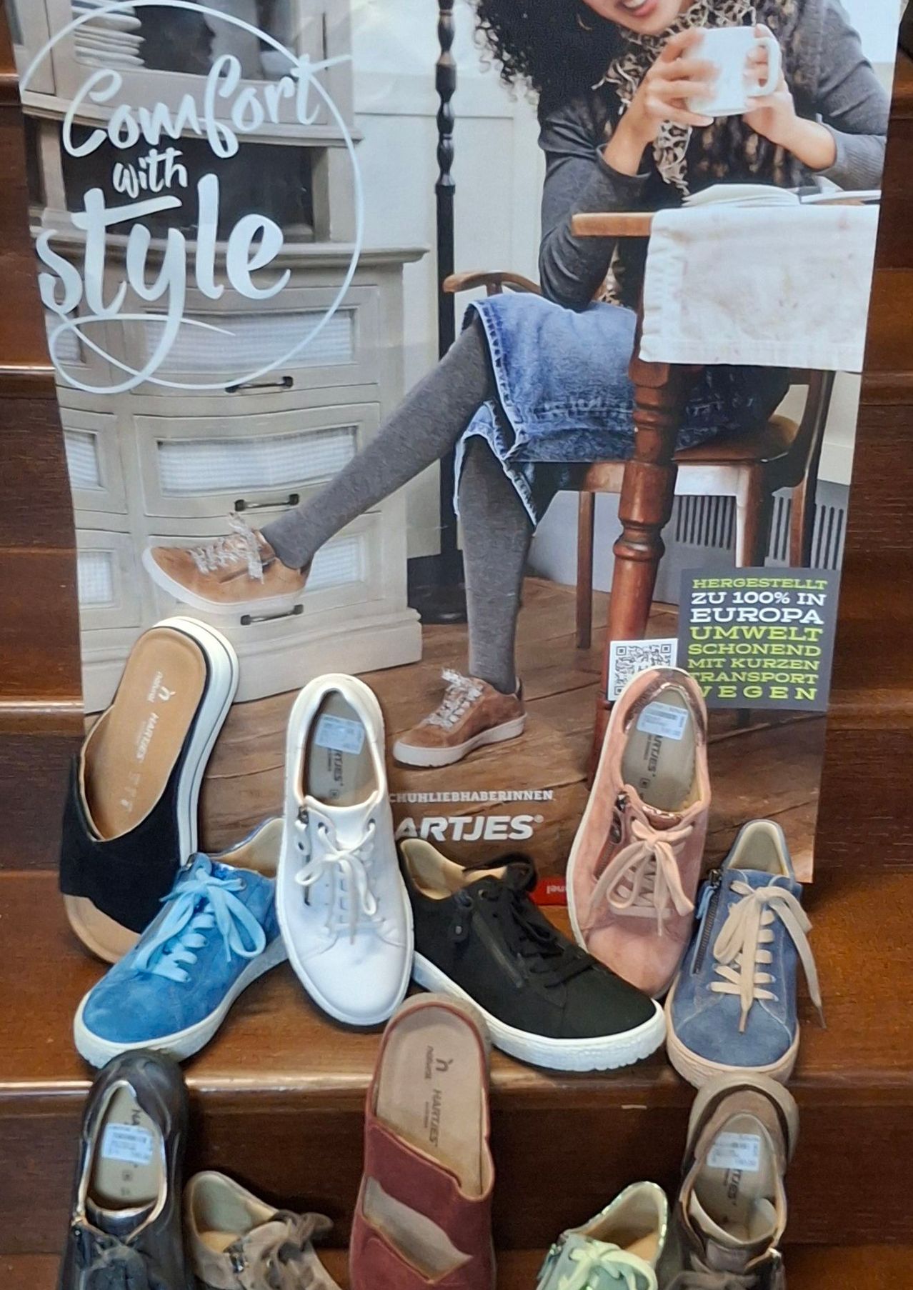 Eine Ausstellung verschiedener stylischer Schuhe, einschließlich schwarzer, weißer, blauer und brauner Paare, auf hölzernen Stufen angeordnet. Eine Person, die Jeansshorts und graue Leggings trägt, sitzt hinter den Schuhen auf einem Stuhl.